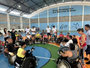 A imagem mostra um grupo de pessoas em cadeiras de rodas reunidas em círculo dentro de um ginásio esportivo, que tem o nome Ginásio Paradesportivo José Belarmino de Souza escrito na parede ao fundo. Os participantes parecem envolvidos em uma atividade ou discussão, com alguns vestindo uniformes esportivos e outros roupas casuais. Ao redor do círculo, há pessoas em pé que podem ser treinadores ou assistentes. O ambiente é iluminado, e o piso da quadra tem marcações coloridas, indicando seu uso para diferentes esportes.